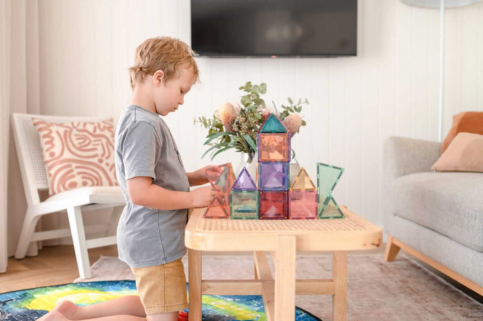 A young boy building a castle structure with pastel magnetic squares and triangles on a wooden table.