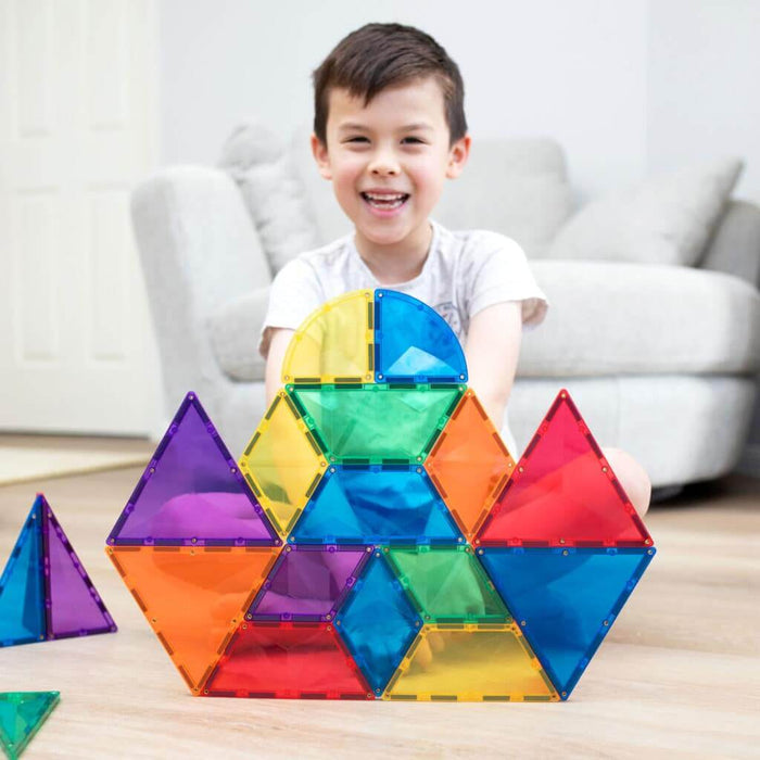 A boy sits behind a colorful structure built with translucent magnetic triangles, diamonds, and hexagons in various colors.
