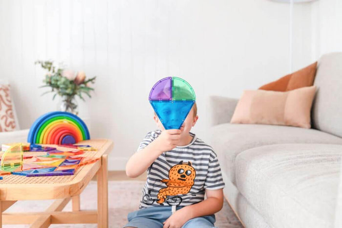 Child holding blue and green translucent magnetic tiles over face with colorful shapes spread on a wooden table.