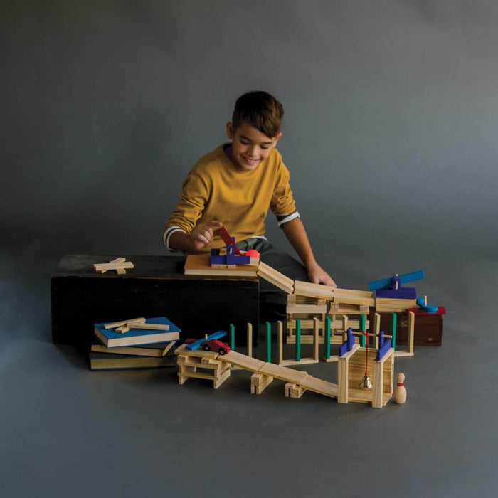 Boy builds a wooden chain reaction course with natural planks, blue and red blocks, a toy car, and a small bell.