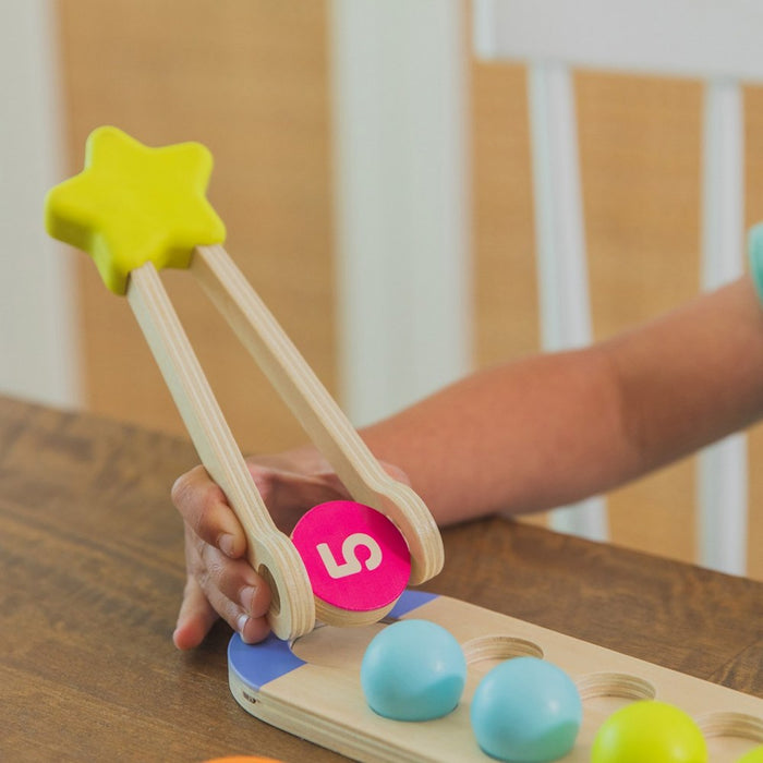 Child using star-shaped wooden tongs to pick up a pink number 5 disc from a wooden tray with colorful spheres.