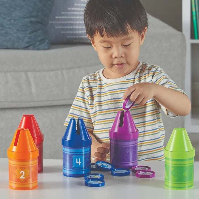 Young boy sorting colored disks into five large plastic crayon banks featuring numbers and shapes.