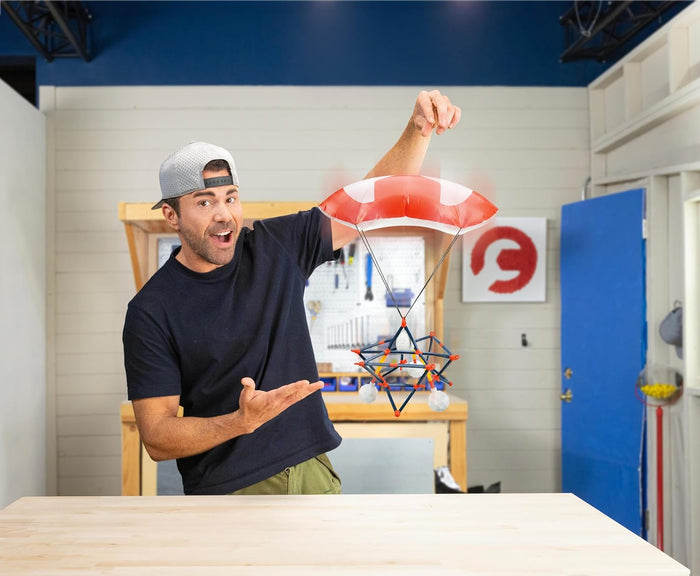 A man in a baseball cap presents an orange and blue geometric frame suspended from a red and white toy parachute.