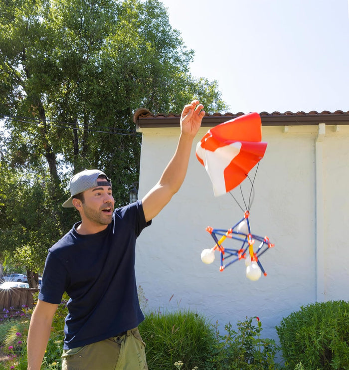A man releases a blue and orange geometric cage with white eggs and an orange and white parachute into the air.