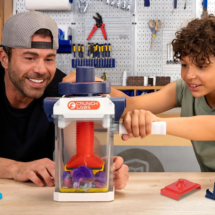 Man and boy use a white and blue toy press with a red screw to squish colorful dough inside a clear plastic chamber.