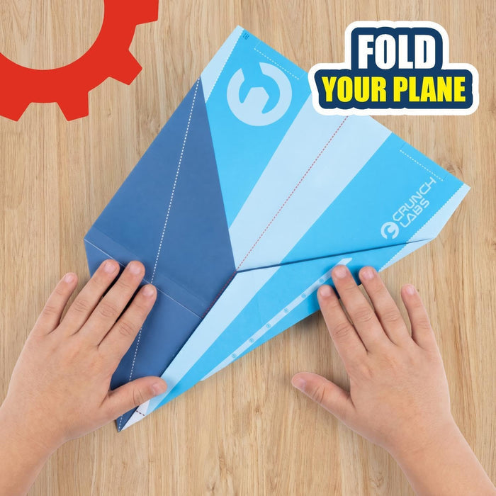 Child's hands folding a blue and white patterned Crunch Labs paper airplane with fold lines on a wooden surface.
