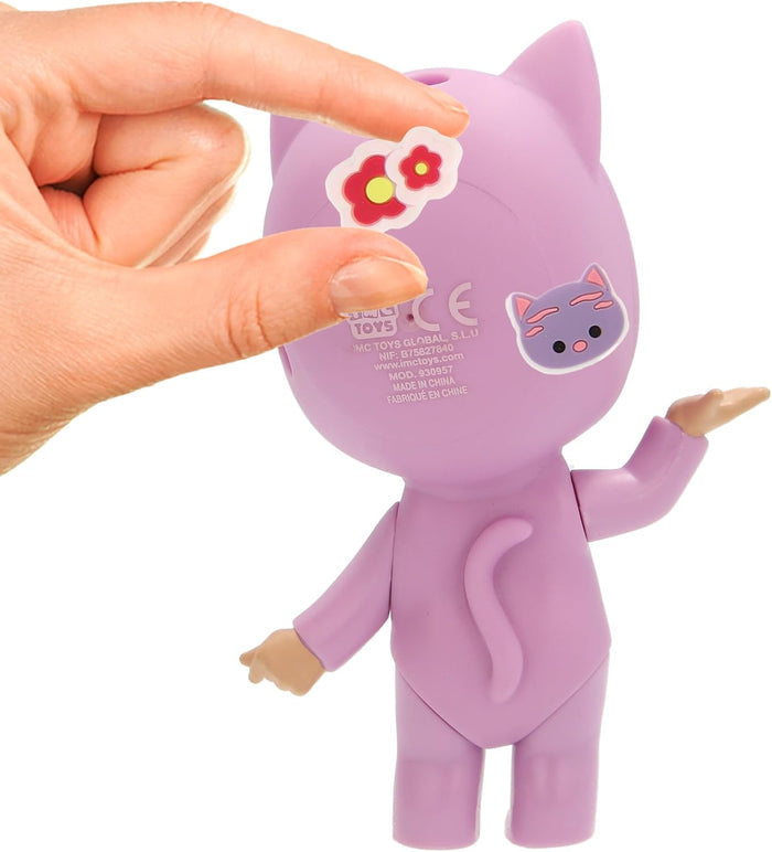 Hand applying a red flower sticker to the back of a purple cat-shaped doll with a tail and tan hands.