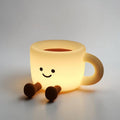 Glowing yellow cup-shaped silicone lamp with a black smiley face and two brown feet on a white surface.
