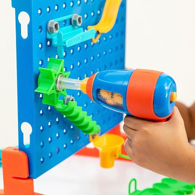 Child's hand uses an orange and blue toy drill to attach a green gear to a blue pegboard with colorful maze parts.