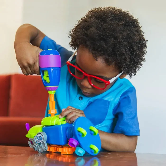 A child wearing red glasses uses a toy drill to build a colorful plastic robot with green bolts and orange gears.