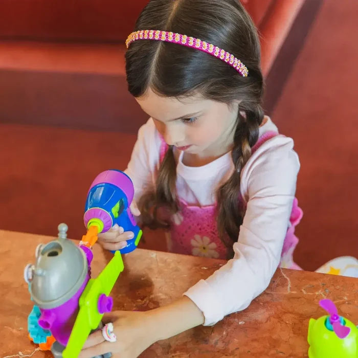 A girl with braided hair uses a colorful toy drill to build a purple and green toy robot on a tabletop.