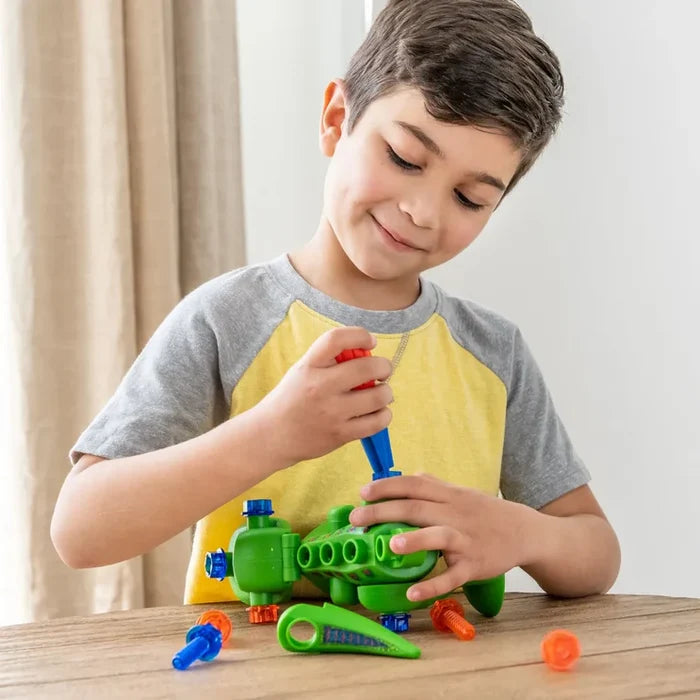 Boy using a toy screwdriver to assemble a green plastic dinosaur toy with colorful bolts on a wooden table.