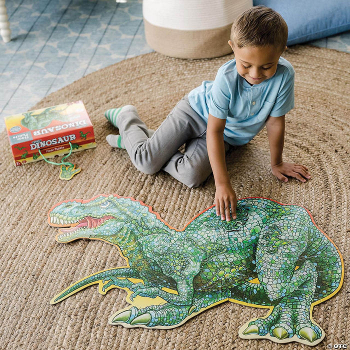 A boy assembles a large, scaly green T-Rex shaped floor puzzle on a rug next to its packaging.