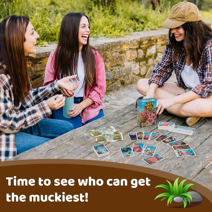 Three friends play a card game with a teal box and pig-themed cards spread across a rustic wooden table outdoors.