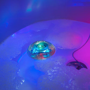 Small, circular light floating in a bubble bath, glowing green and blue with pink and purple reflections on the water.