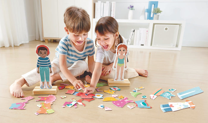 Children playing with stand-up magnetic dolls and colorful clothing puzzle pieces on a wooden floor.