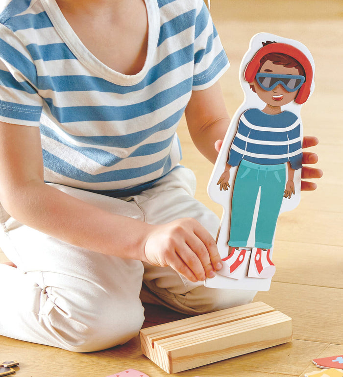 Child outfits a magnetic boy doll with a striped sweater and red beanie on a wooden stand.