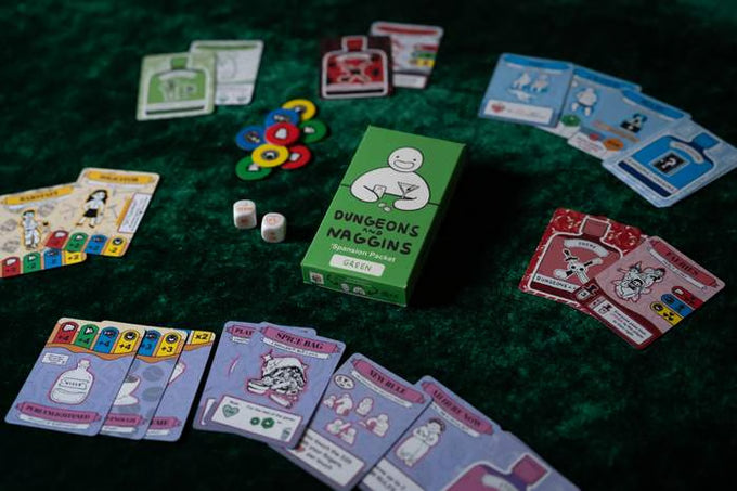 Green game box with a white cartoon figure on green felt, surrounded by playing cards, dice, and colorful tokens.