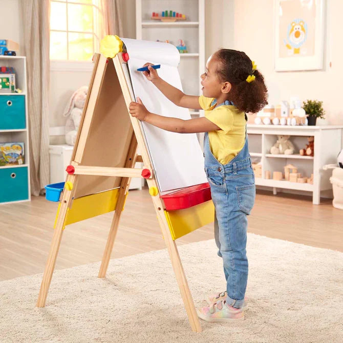 Young girl drawing with a blue marker on a white paper pad roll attached to a wooden standing easel.