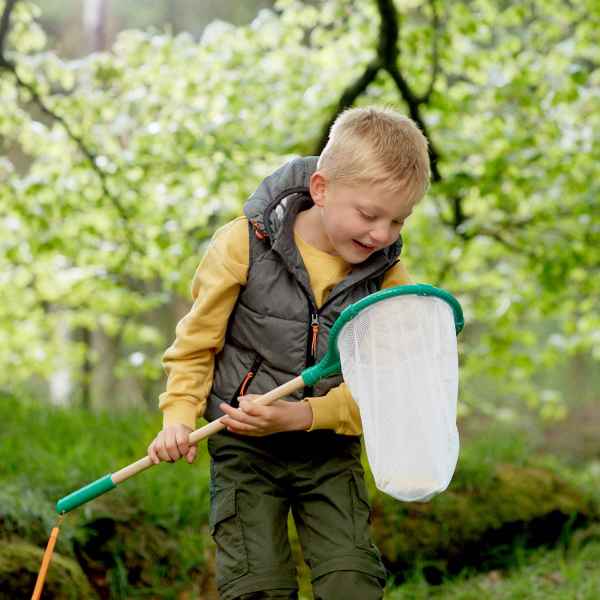 A child holding a white mesh net with a wooden handle and green trim in a lush green outdoor setting.