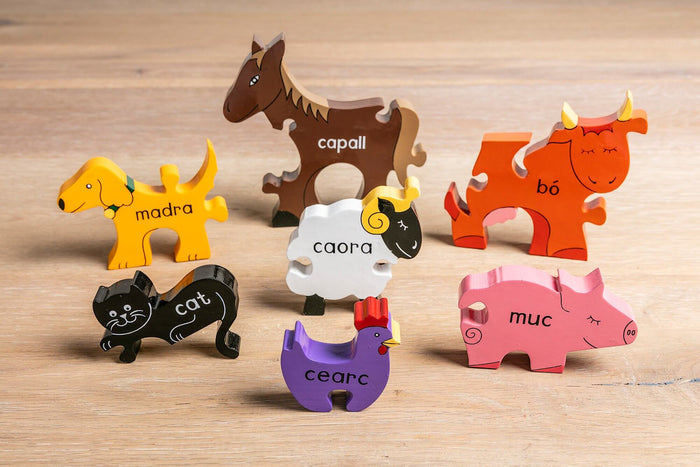 Seven colorful wooden animal puzzle pieces labeled with Irish words stand on a light wood surface.