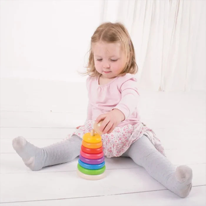 Toddler sitting on floor stacking a wooden toy with seven colorful rainbow rings and a natural wood ball topper.