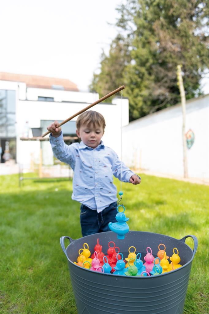 A child catches colorful plastic ducks with head loops from a gray tub using a wooden toy rod on a green lawn.