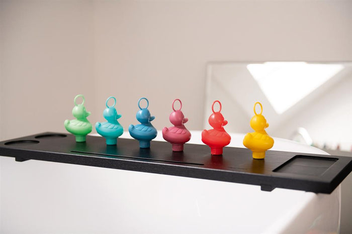 Six rubber ducks in rainbow colors with head rings lined up on a black bath caddy over a white tub.