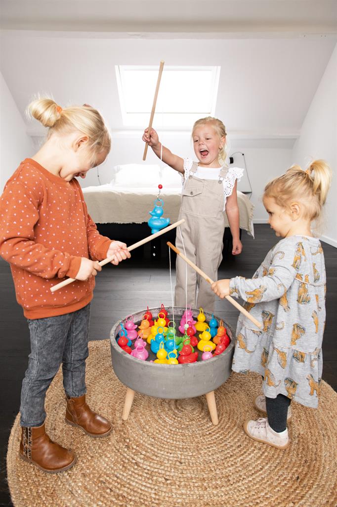 Three girls use wooden fishing rods to hook colorful plastic ducks from a circular grey basin on a woven rug.