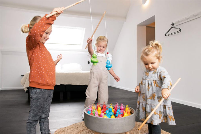Three children use wooden rods to catch colorful red, yellow, and blue plastic ducks from a round grey basin.