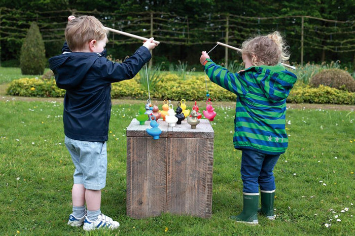 Children use wooden rods to catch colorful plastic ducks with head loops from a wooden crate in a garden.