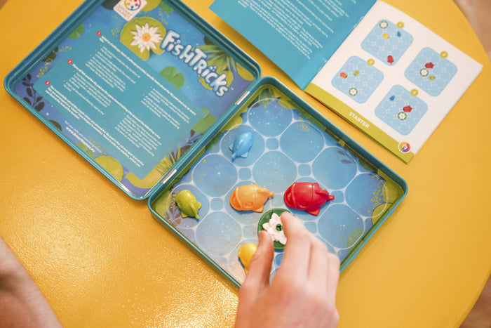 Hand playing FishTricks game in a blue tin with colorful magnetic fish, a lily pad, and a challenge booklet.