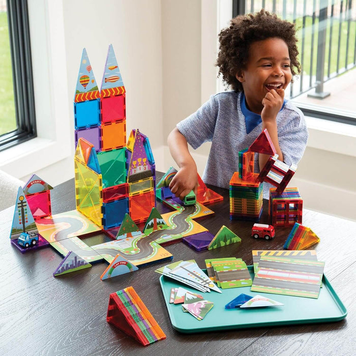 Boy playing with colorful magnetic tiles featuring road graphics, building structures, and small toy vehicles.