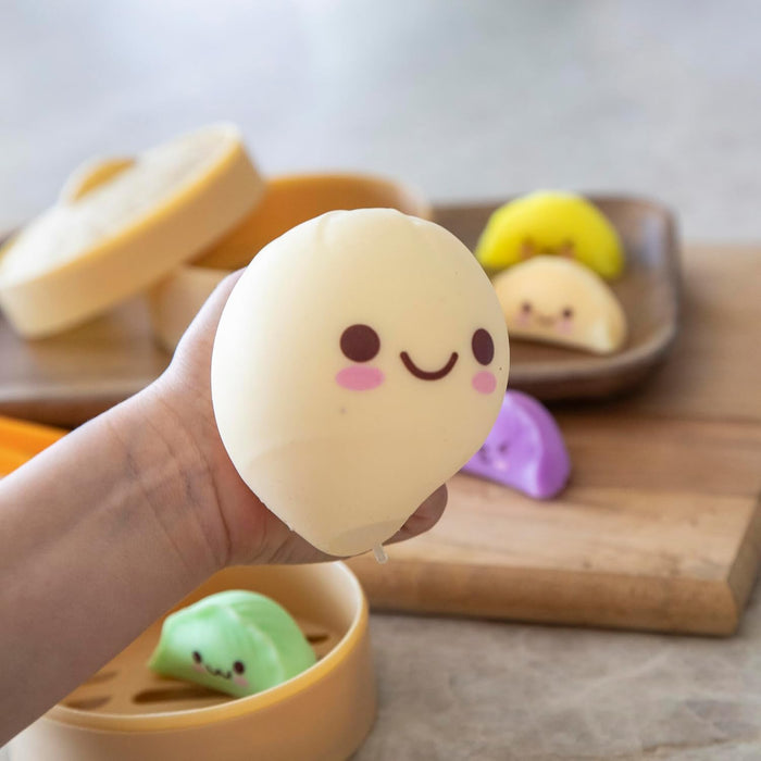 A hand holds a cream-colored squishy bun toy with a happy face. Colorful dumpling toys and toy steamers sit nearby.