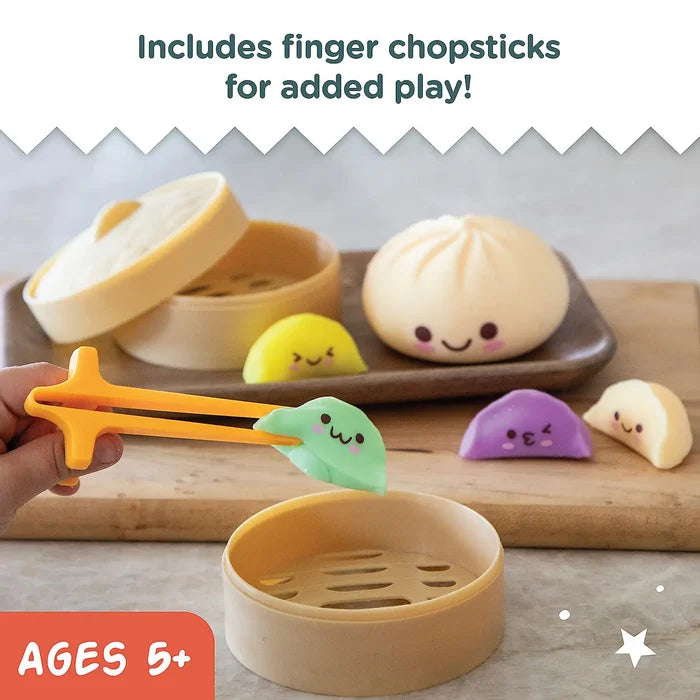 Hand using orange finger chopsticks to pick up a green squishy dumpling toy among colorful buns and tan steamer baskets.