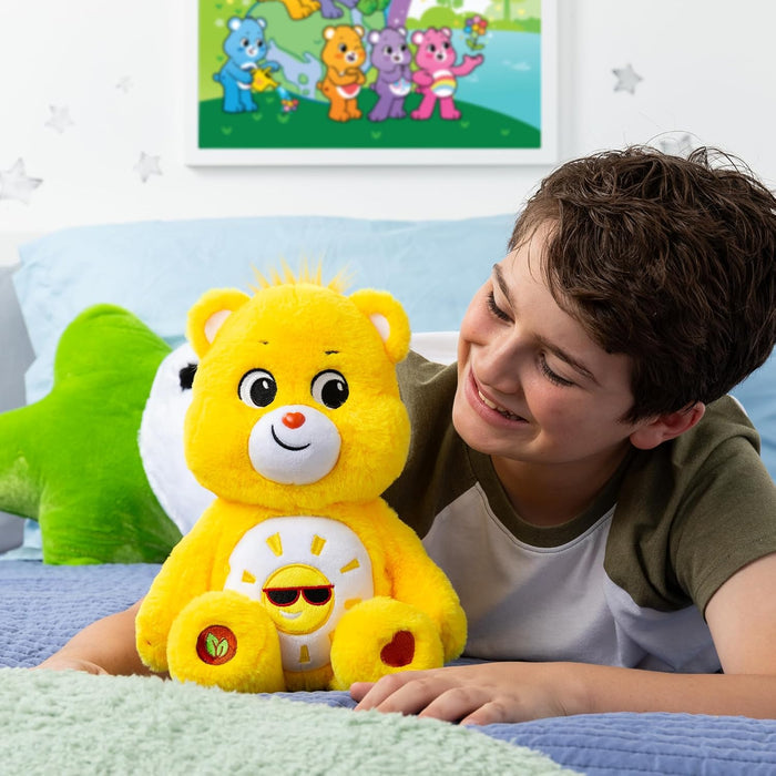 Bright yellow plush Care Bear with a sun belly badge, sitting next to a smiling boy on a bed.