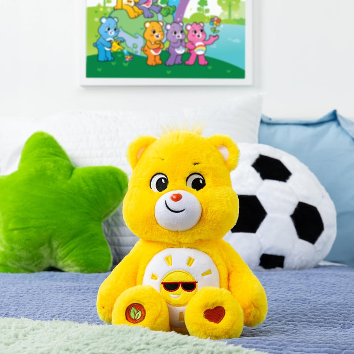 Bright yellow plush bear with a sun wearing sunglasses on its belly, sitting on a bed with star and soccer pillows.