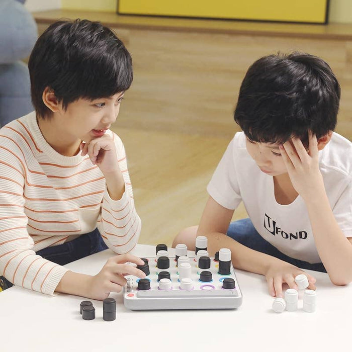 Two boys play a strategy game with stackable black and white cylinders on a white square board with colorful ring lights.