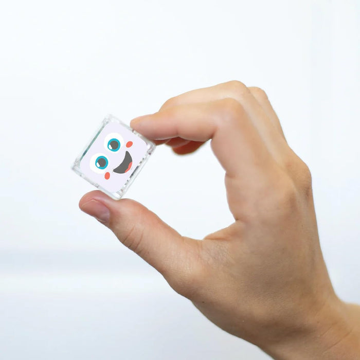 A hand holds a small, clear square cube featuring a smiling white cartoon face with blue eyes and pink cheeks.