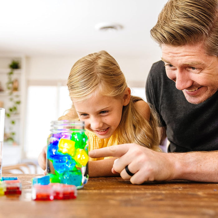 A man and young girl look at glowing green, yellow, and blue cubes inside a glass jar of water on a wooden table.