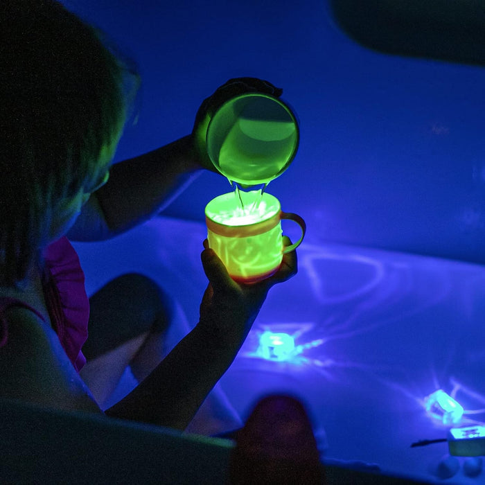 Child pours water into a glowing green mug in a dark bath with small light-up cubes shining bright underwater.
