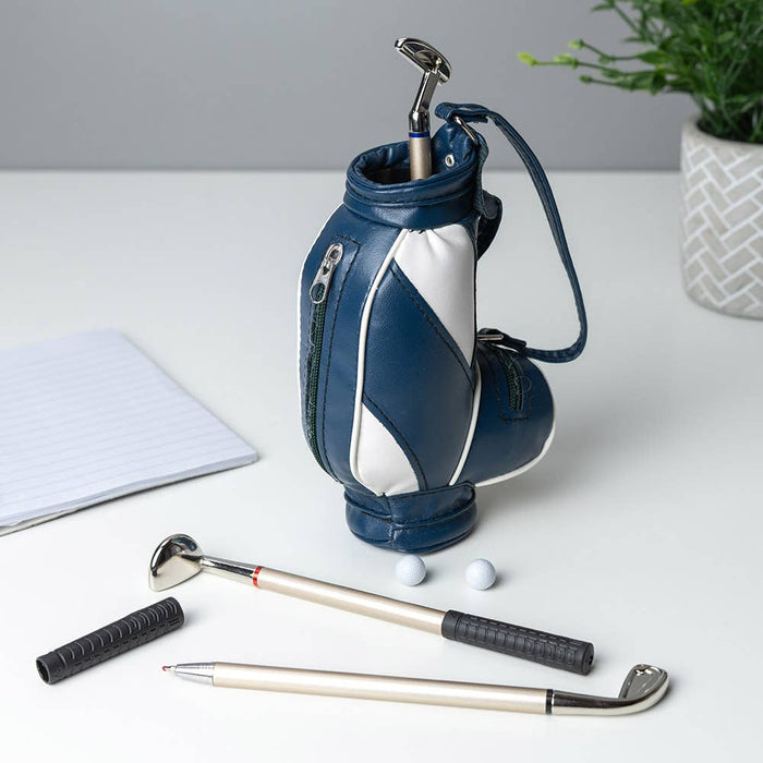 Miniature blue and white golf bag holding golf club-shaped pens and two tiny white balls on a white desk.