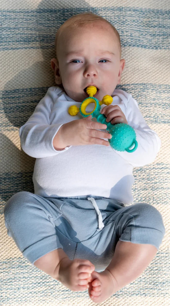 Baby lying on a rug chewing on a teal and yellow textured teether toy with a ring and knobby ball.