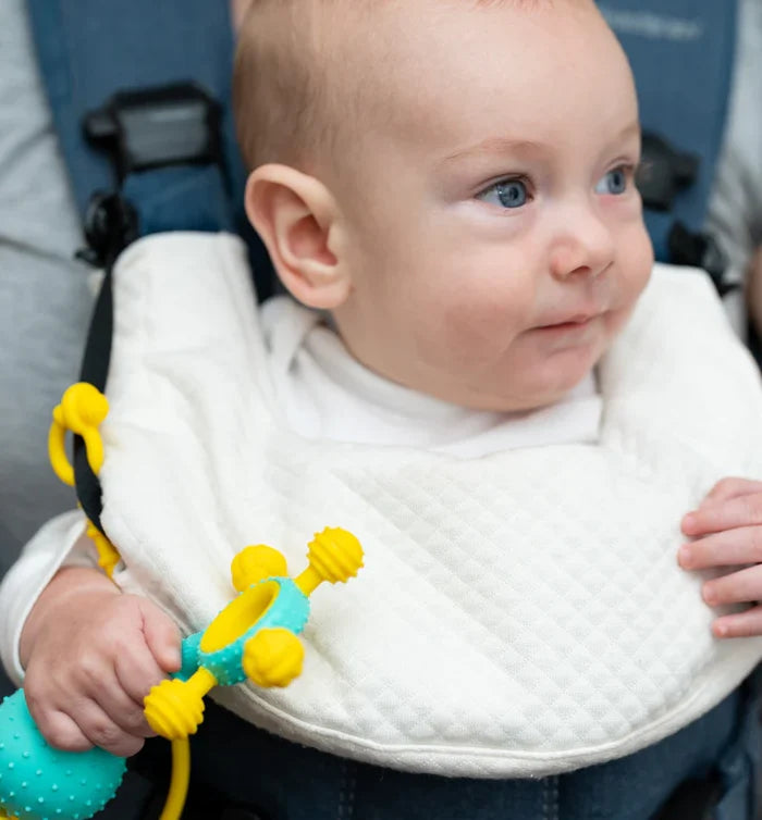 A baby in a carrier holds a colorful textured silicone teething toy with yellow nubs and a teal ring.