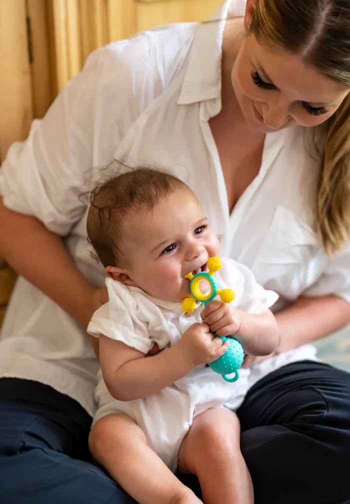 Baby chewing a teal and yellow silicone teething toy with a bumpy ball base and a multi-knobbed ring top.
