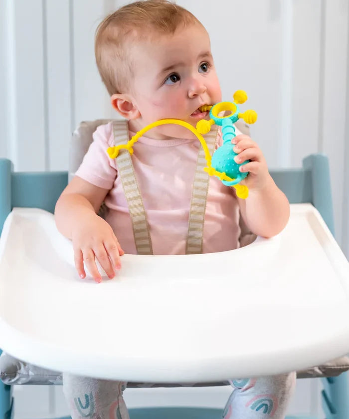 A baby in a high chair holding a teal and yellow silicone teething toy with textured surfaces and a flexible tether.