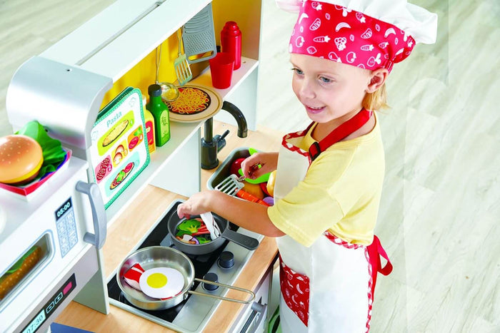 Child in a red chef hat plays with a white toy kitchen featuring a stovetop, microwave, and play food.