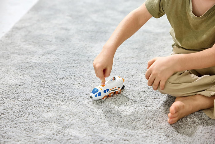 Child playing on a grey rug with a white toy shuttle train featuring blue windows and orange wheels.