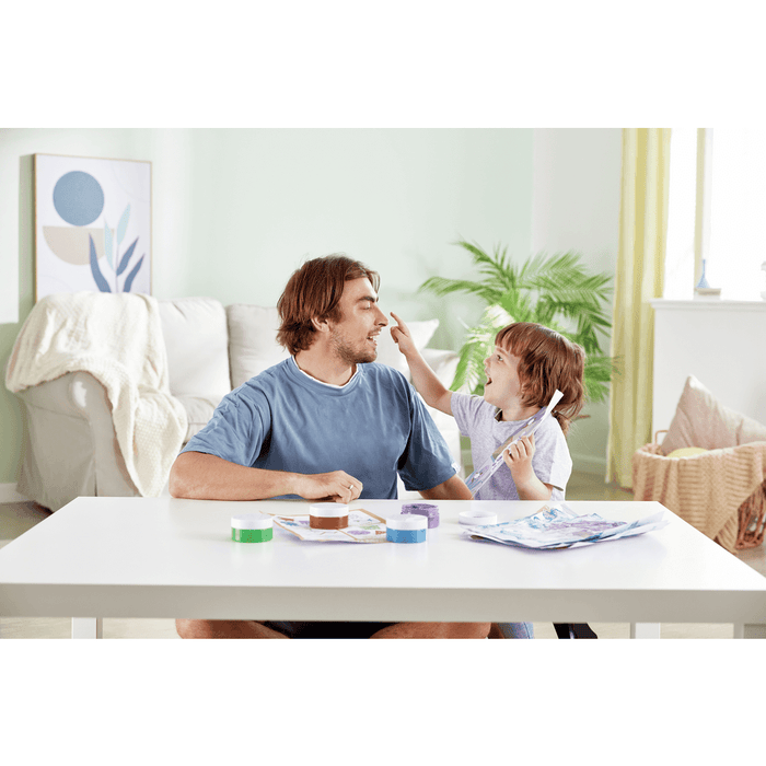 A child touches a man's nose while playing with small, colorful finger paint pots and paper on a white table.