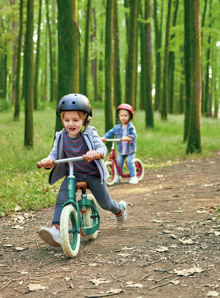 Two toddlers ride green and red no-pedal balance bikes with white tires on a wooded dirt path. Both wear safety helmets.